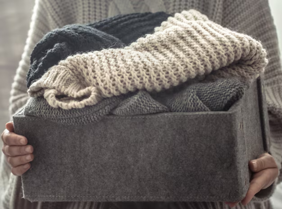 A person holding a felt box filled with cozy knitted sweaters in neutral tones.