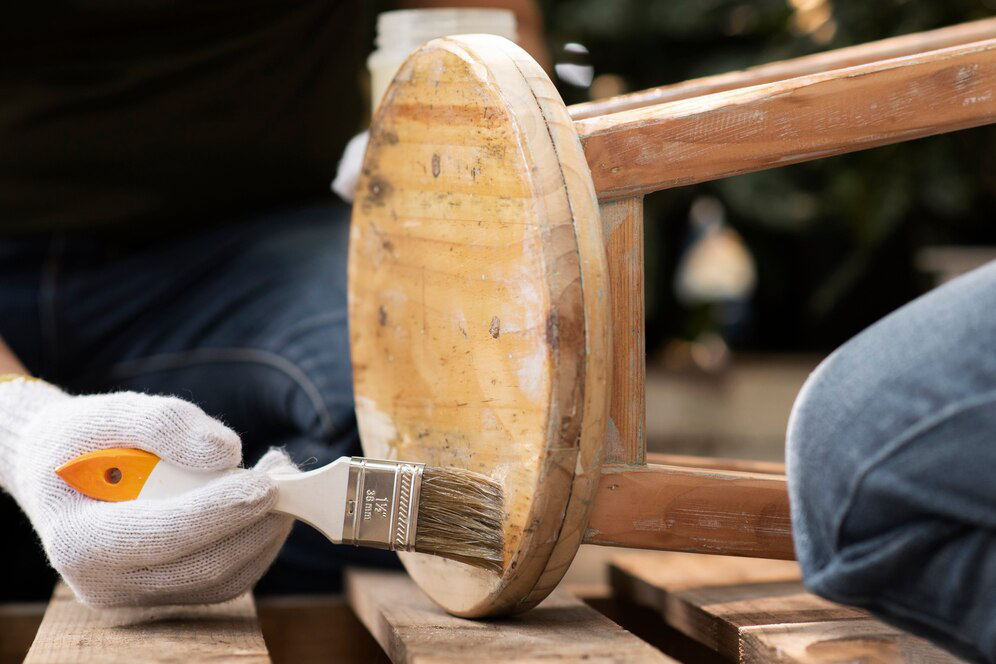 A person with gloved hands paints the bottom of a wooden stool using a brush.