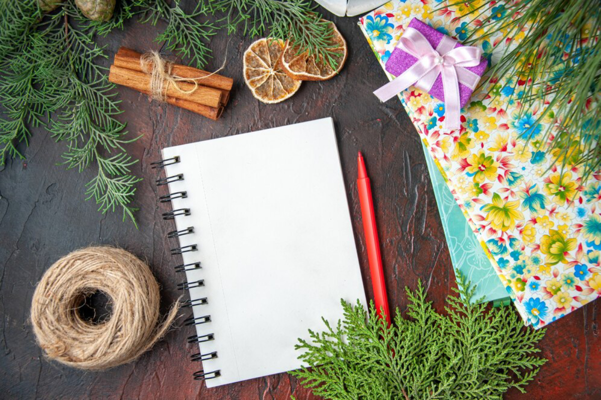 A blank notebook, red pen, twine, cinnamon sticks, dried oranges, and floral paper.