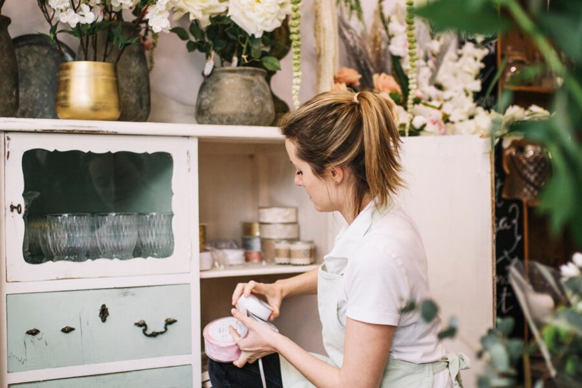 A woman in an apron organizes ribbon spools inside a vintage cabinet surrounded by beautiful flower arrangements and decorative pots.