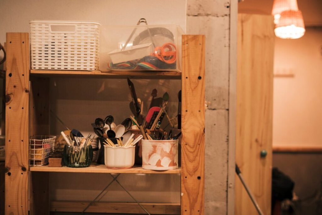 A wooden shelf filled with art supplies, including brushes, spoons, and containers, set against a softly lit wall.